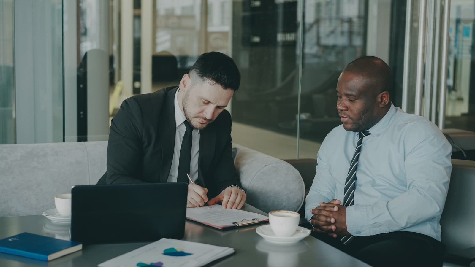 Two businessmen at an office table, one signing documents as the other watches