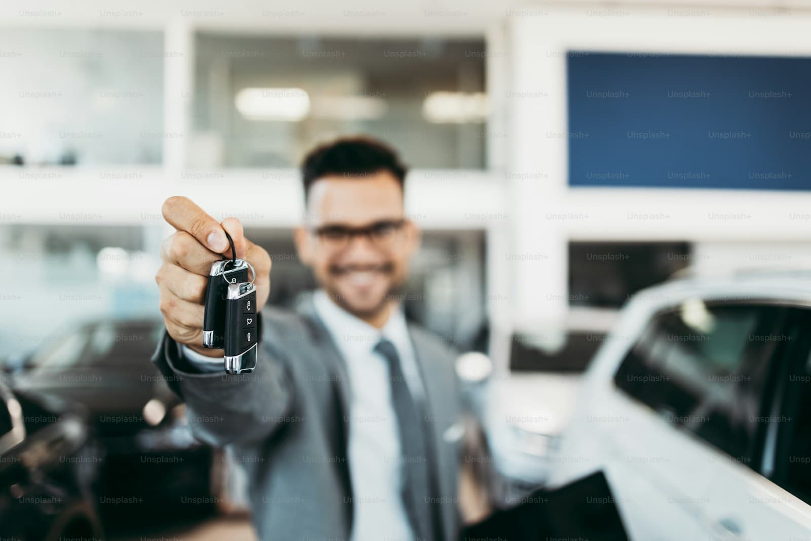 Smiling car salesman in suit holding out car keys at automotive dealership showroom