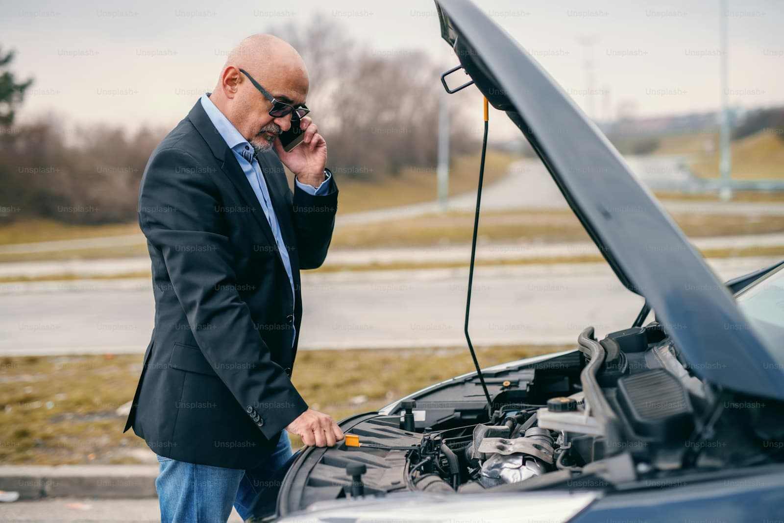Man in a suit phoning for assistance beside his car with the hood raised