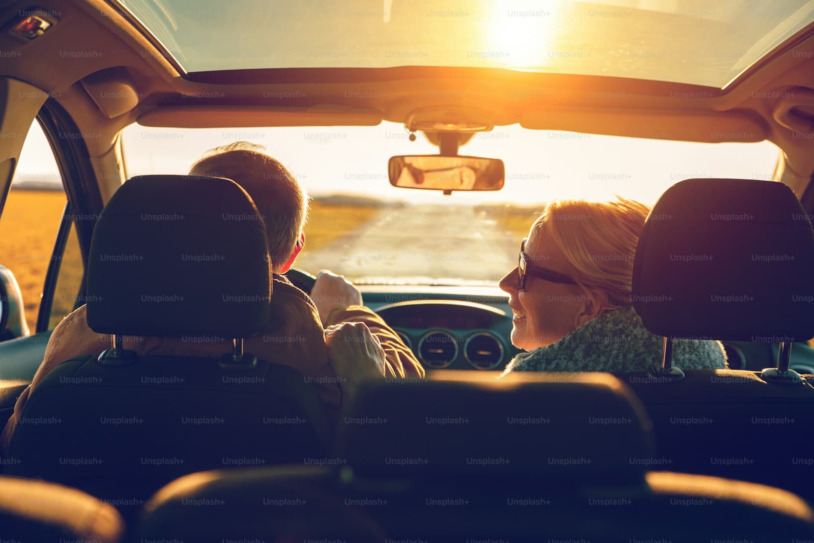 Two people driving toward a bright sunset with golden light through the windshield