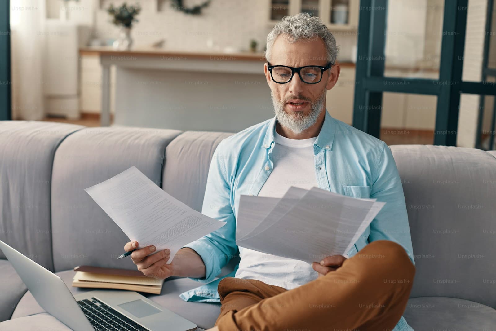 Mature man with glasses reading papers while working on laptop at home on gray couch