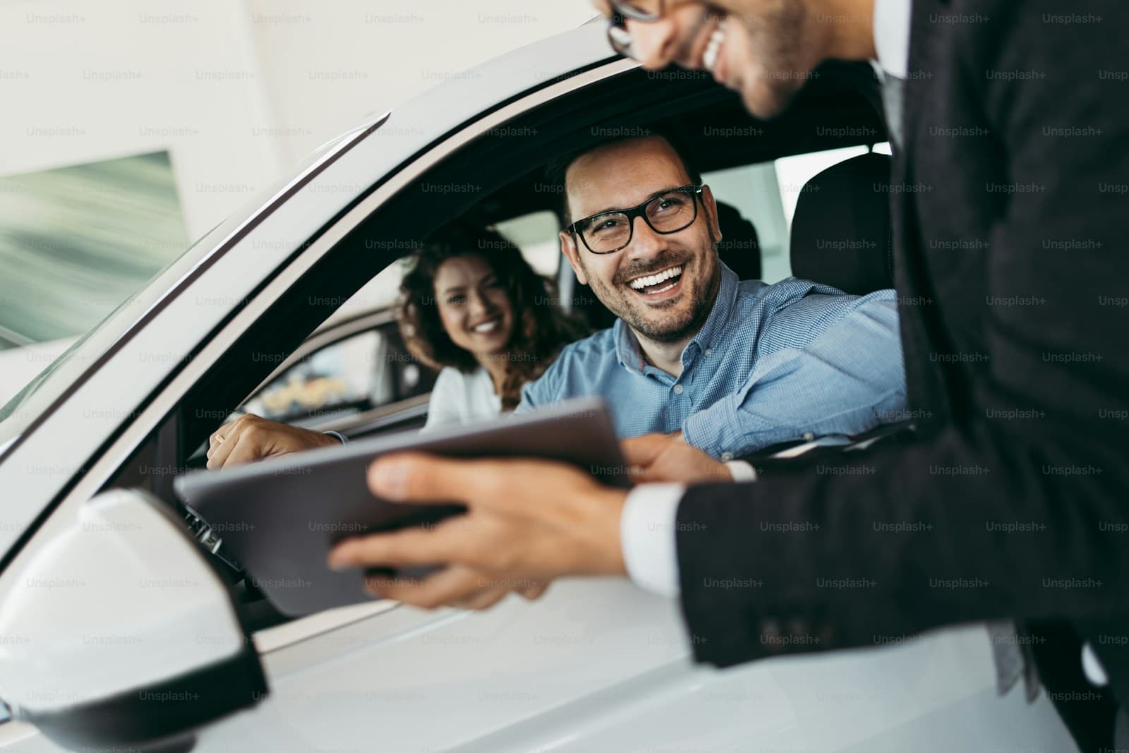 Smiling man in glasses in the driver's seat as someone leans in through the open window