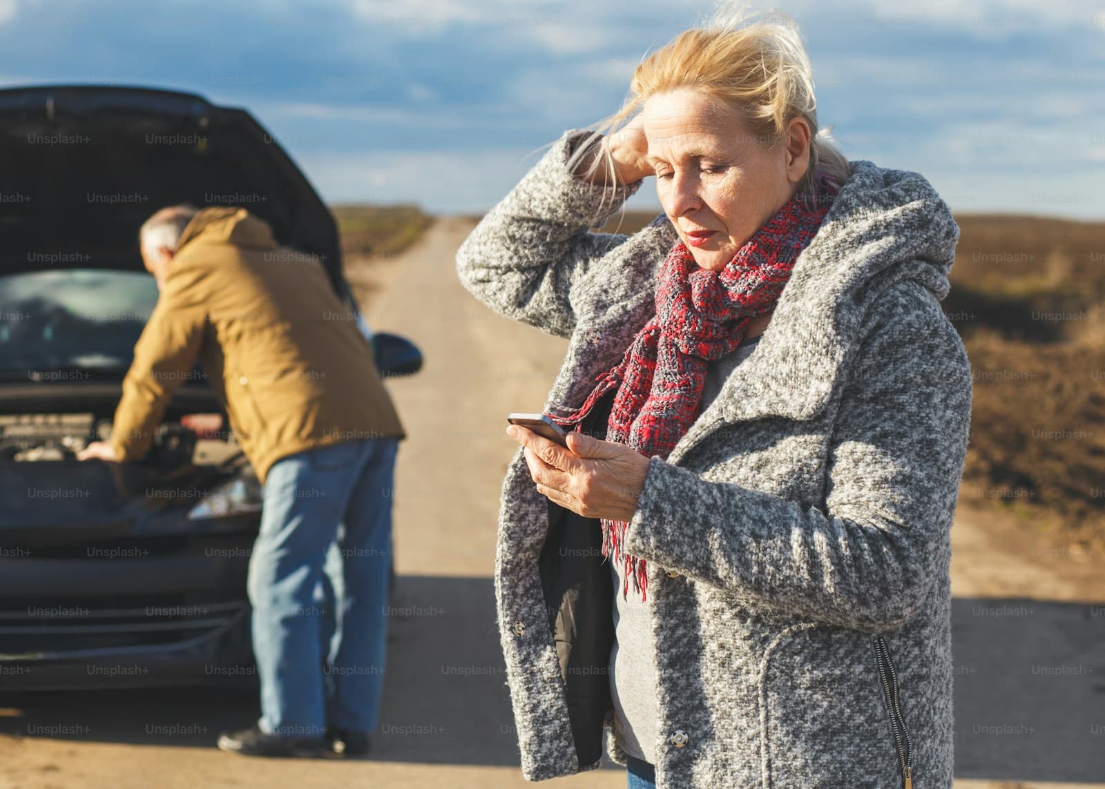 Woman in a gray coat and red scarf calling for help as a man checks under the hood