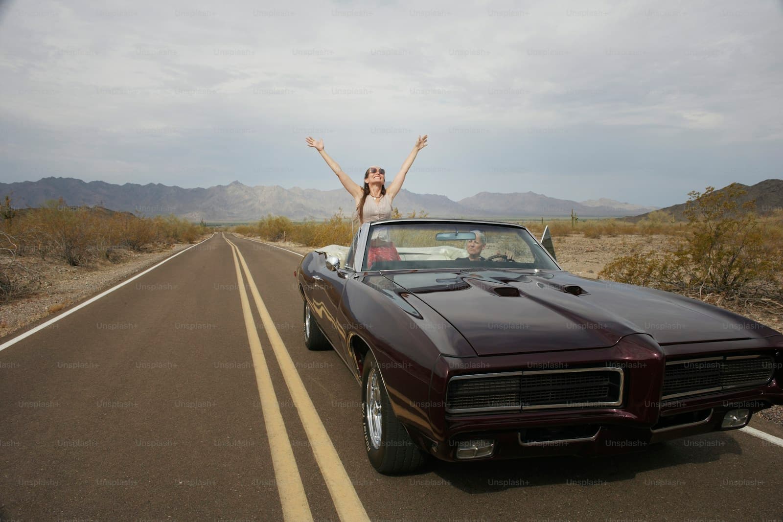 Woman with arms raised standing through sunroof of vintage convertible muscle car on empty desert highway