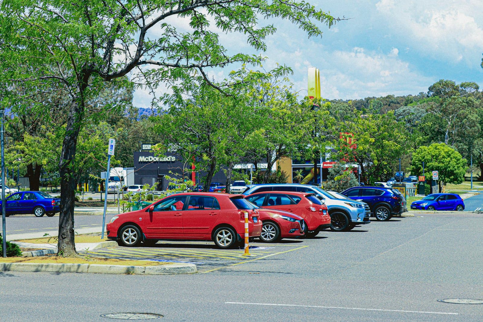 Cars parked in a lot with red sedan in foreground, green trees and hills in background under cloudy sky