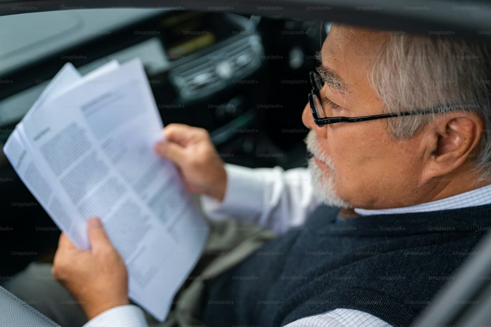 Man in glasses and a dark sweater reading paperwork in the driver's seat of his car