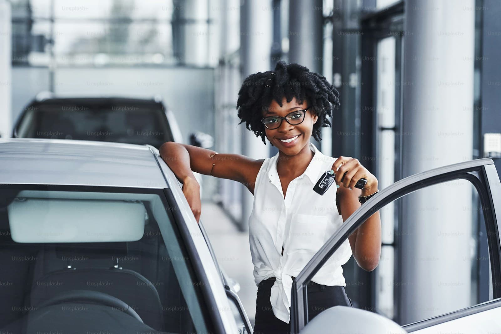 Happy Black woman with dreadlocks holding car keys next to white car in dealership showroom