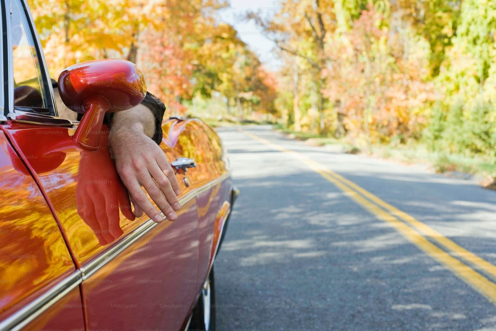 Driver's arm out the window of a red car on a tree-lined road in golden autumn foliage