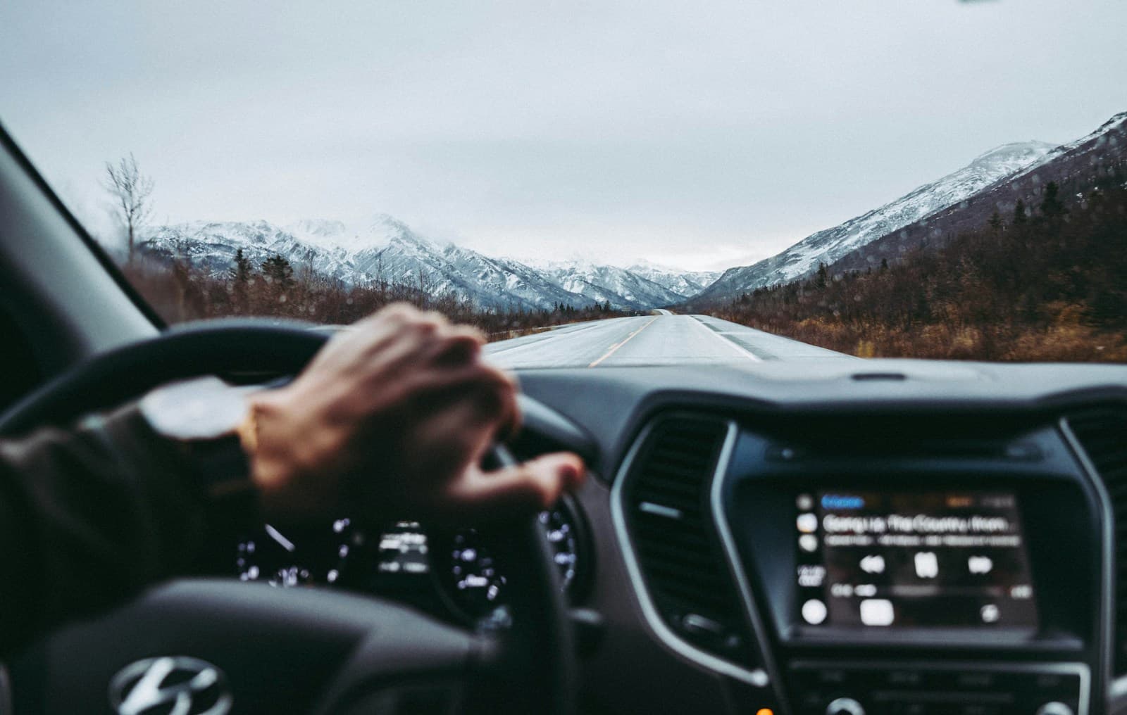 Driver on a straight road toward snow-capped peaks with autumn hillsides on either side